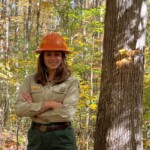 Kelly Corbine in Forester uniform and orange hard hat standing with her hands crossed in a forest