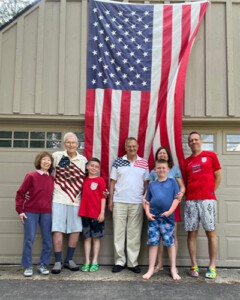 George Treier with his wife Ronna, his brother and grandchildren standing infront of a big flag of United States of America
